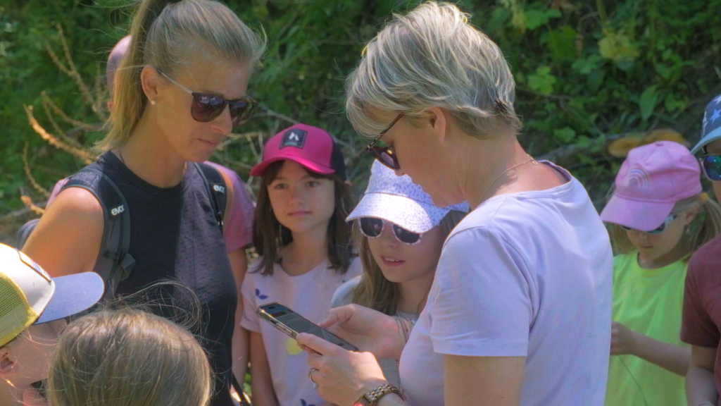 Gruppe von Erwachsenen und Kindern auf dem Eichhörnchenweg in Bürchen, eine Frau schaut auf ihr Smartphone, während die Kinder gespannt zusehen.