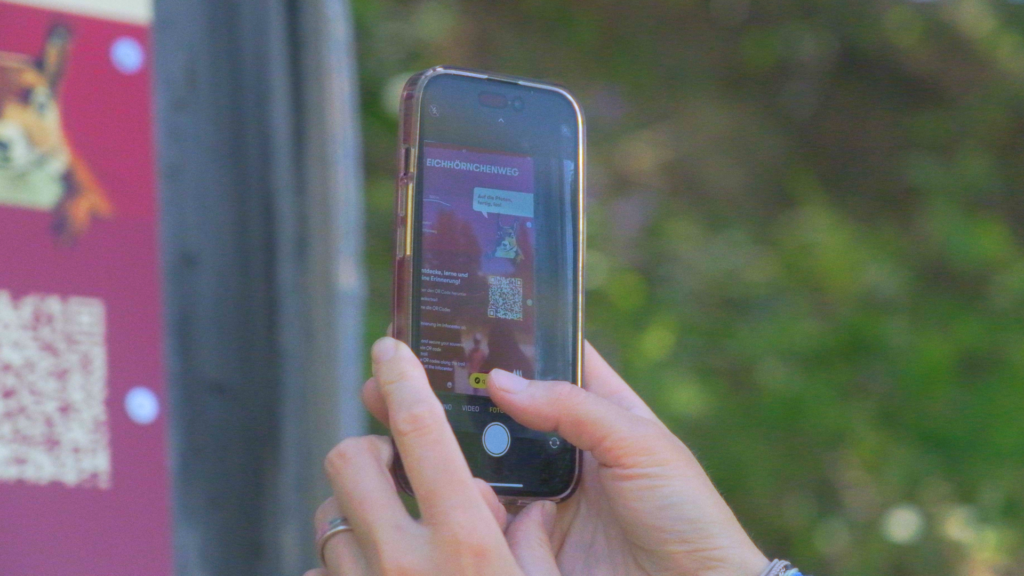 Nahaufnahme eines Smartphones, das einen QR-Code am Eichhörnchenweg scannt, um ein Rätsel in der Moosalp-App zu starten.