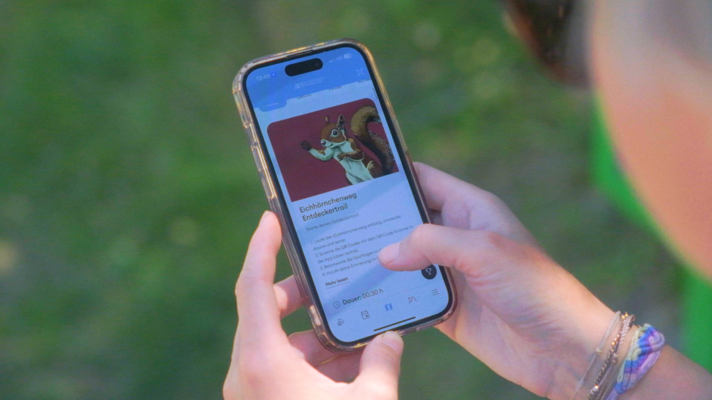 Person hält ein Smartphone in der Hand, auf dem die Moosalp-App mit Informationen zum Eichhörnchenweg Entdeckertrail angezeigt wird.
