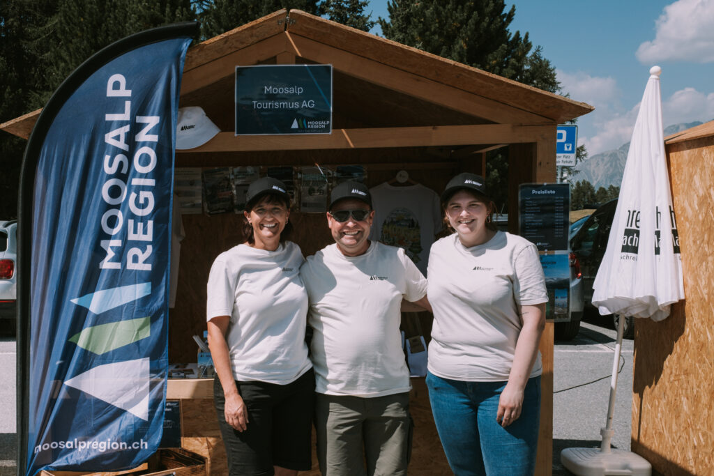 Drei Mitarbeitende der Moosalp Tourismus AG stehen lachend vor einem Infostand mit Banner „Moosalp Region“.